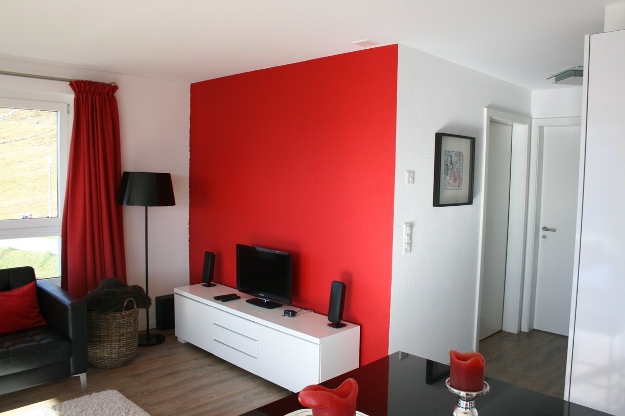 Un salon moderne avec un mur d’accent rouge, un meuble de rangement blanc pour l’équipement audiovisuel, un canapé noir et un objet décoratif rouge posé sur la table.