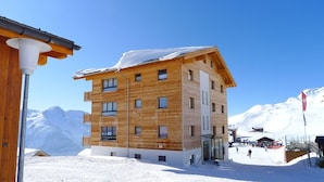 A multi-story wooden building with a flat roof, likely a hotel or resort, situated in a snowy mountainous area with clear skies. - Bella Vista (Fiesch)