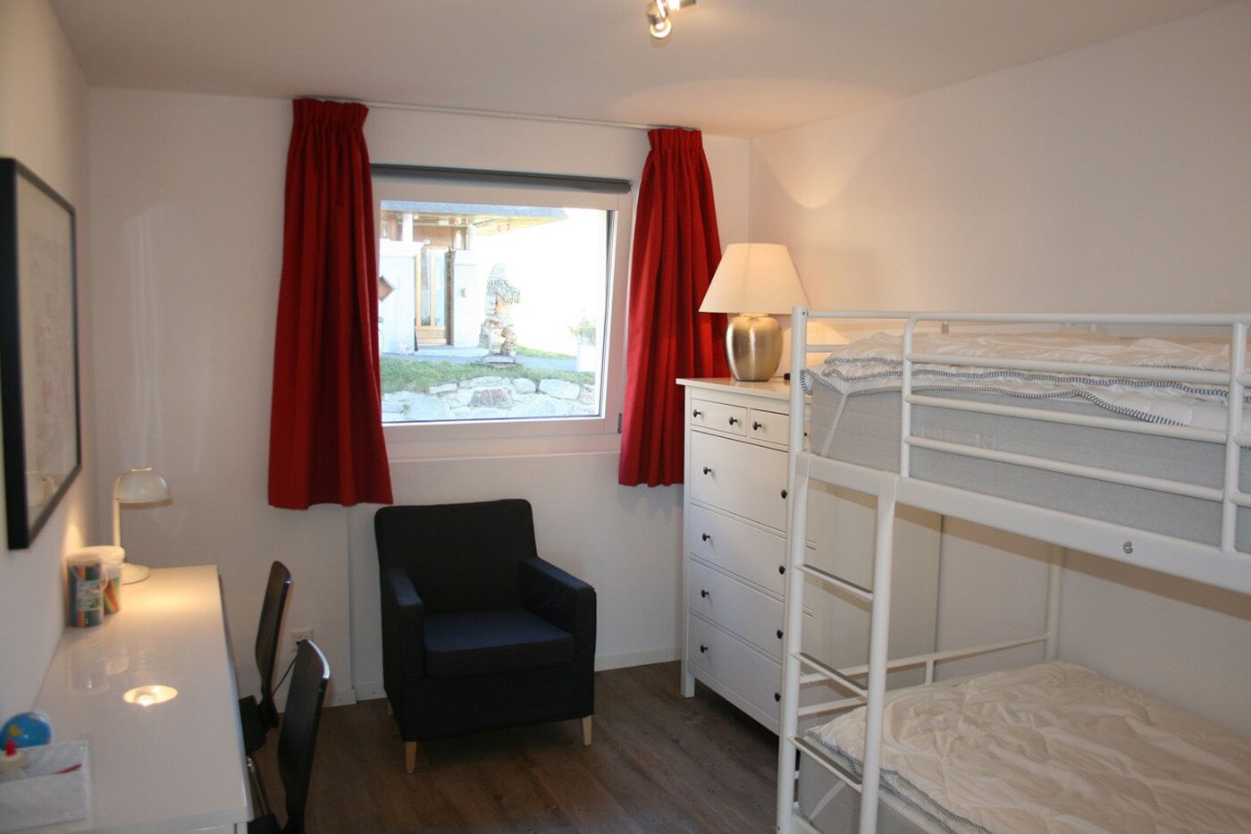 A small, tidy room with a bunk bed, a desk with a lamp, a chair, and a window with red curtains.