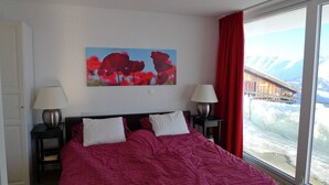A bedroom with a large bed, two bedside lamps, a painting of red flowers, and a window with a view of mountains. - Bella Vista (Fiesch)