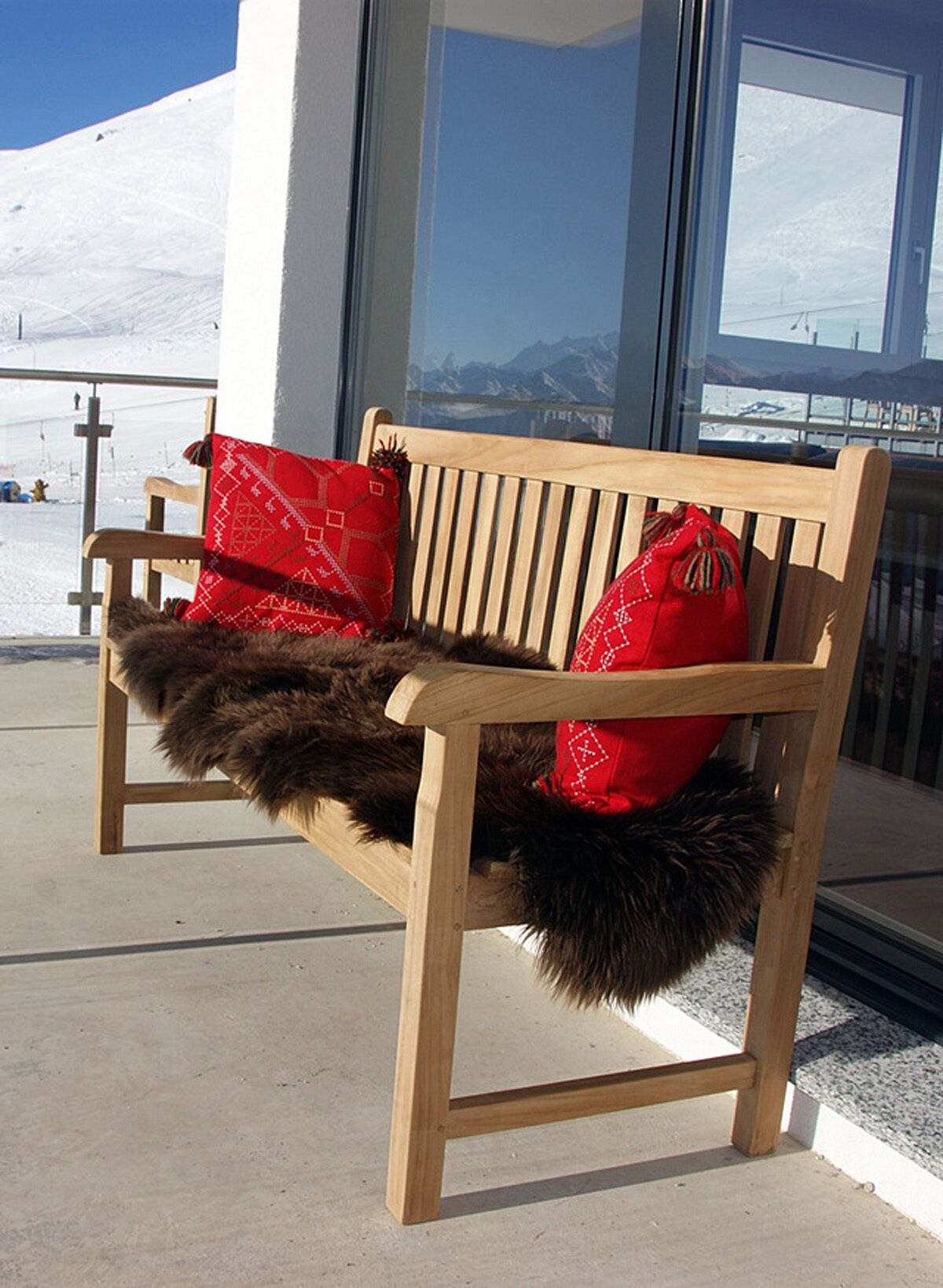 A wooden bench with red cushions and a fur throw, placed outside a building with a view of a snowy mountain.