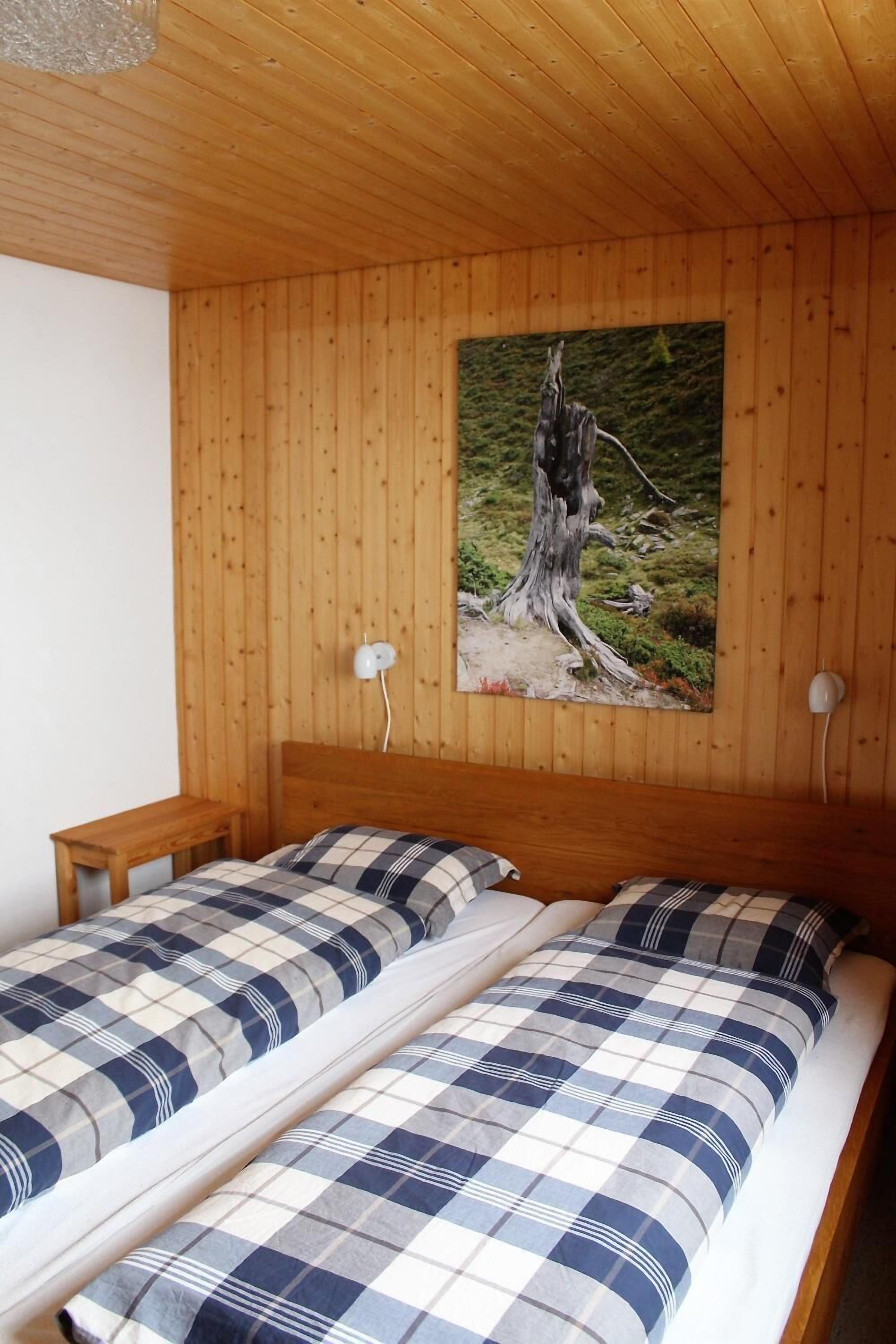 A room with two beds, wooden walls, a painting of a tree, and a white lamp.