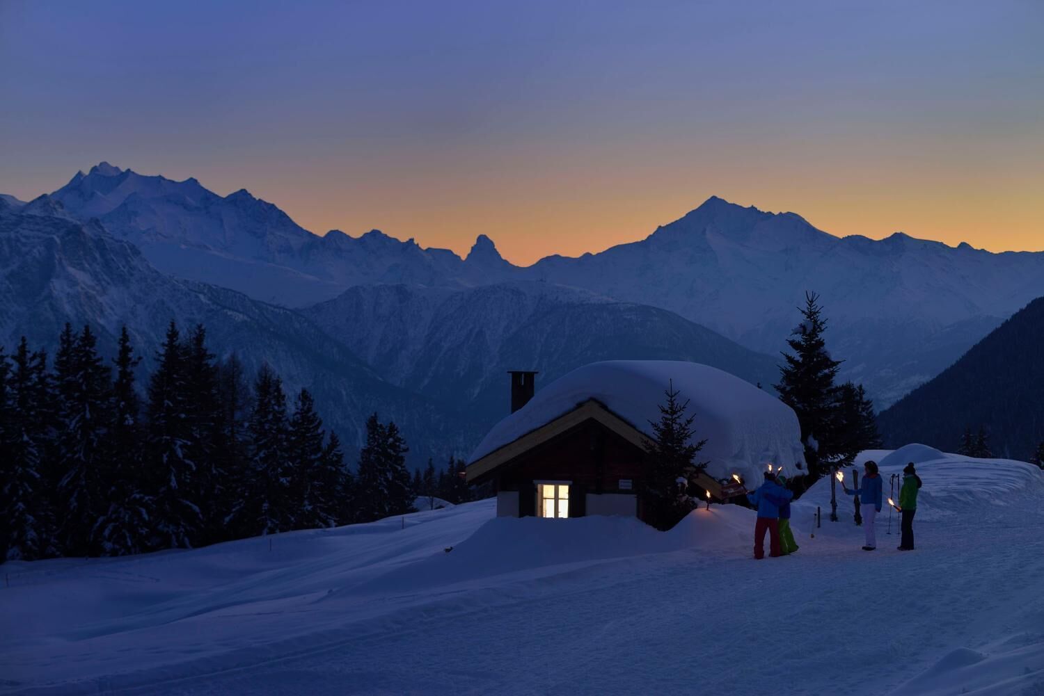 A snow-covered mountain landscape at dusk with a cabin and skiers.
