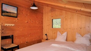 A wooden cabin room with a bed, a chair, a nightstand, a lamp, a window, and a picture on the wall. - Carpe Diem 2 (Fiesch)