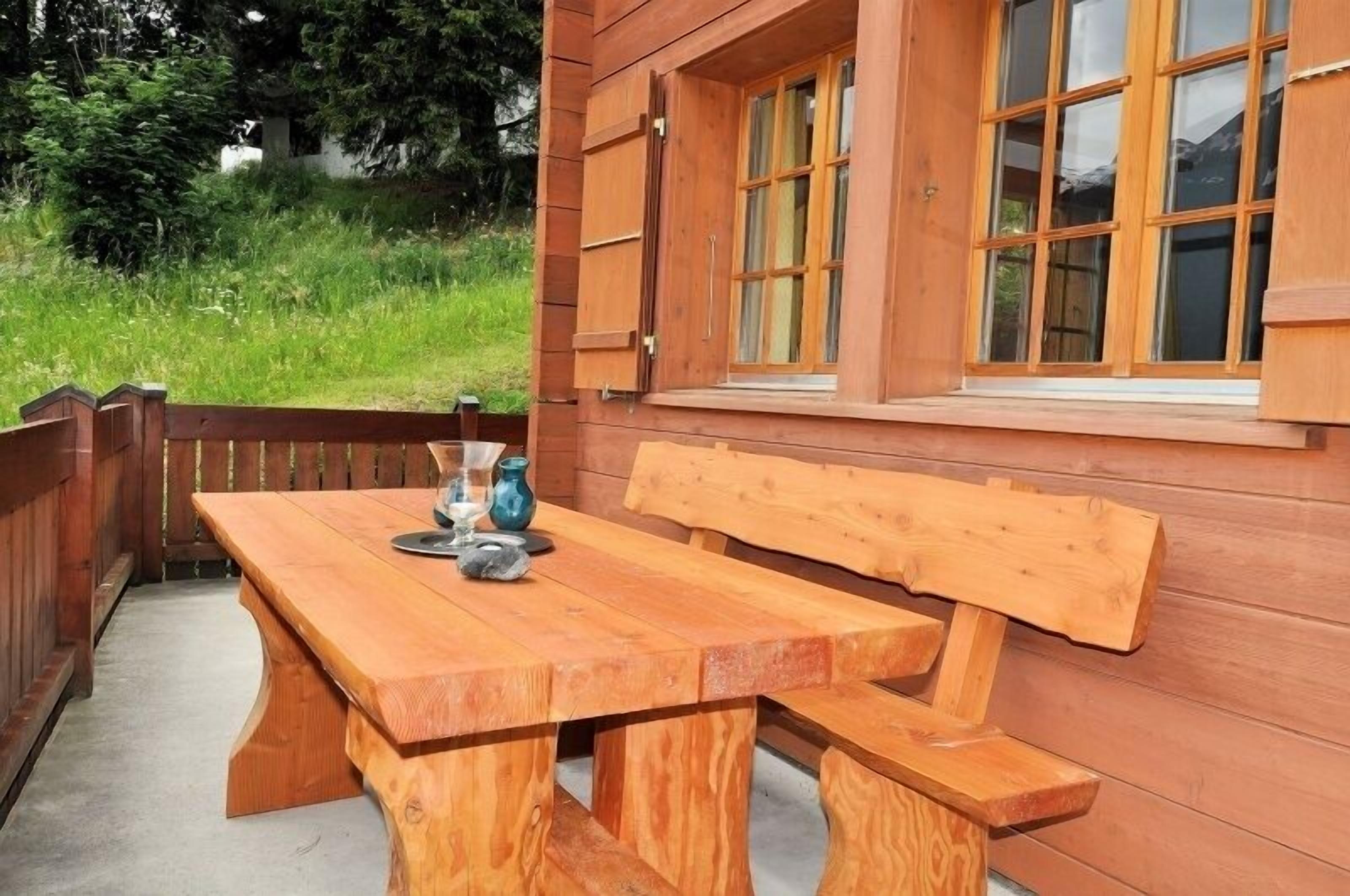 A wooden outdoor seating area with a bench and table, surrounded by a wooden fence and a window with multiple panes.