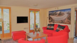 A living room with a red sofa, a wooden coffee table, and a painting of a mountain landscape. - De Hooghe Stee (Fiesch)