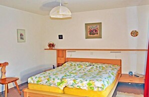 A bedroom with a wooden bed, a bedside table, a chair, and a hanging light fixture. - Dorothy Sunshine (Fiesch)