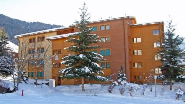 Un edificio de varios pisos con fachada de ladrillo y numerosas ventanas, rodeado de árboles cubiertos de nieve y un telón de fondo montañoso.