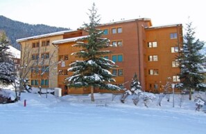 A multi-story building with a brick facade and multiple windows, surrounded by snow-covered trees and a mountainous backdrop. - Dorothy Sunshine (Fiesch)