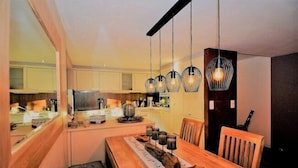 A modern dining area with a wooden table, hanging pendant lights, and a view of the kitchen in the background. - Edelweiss Eden (Fiesch)