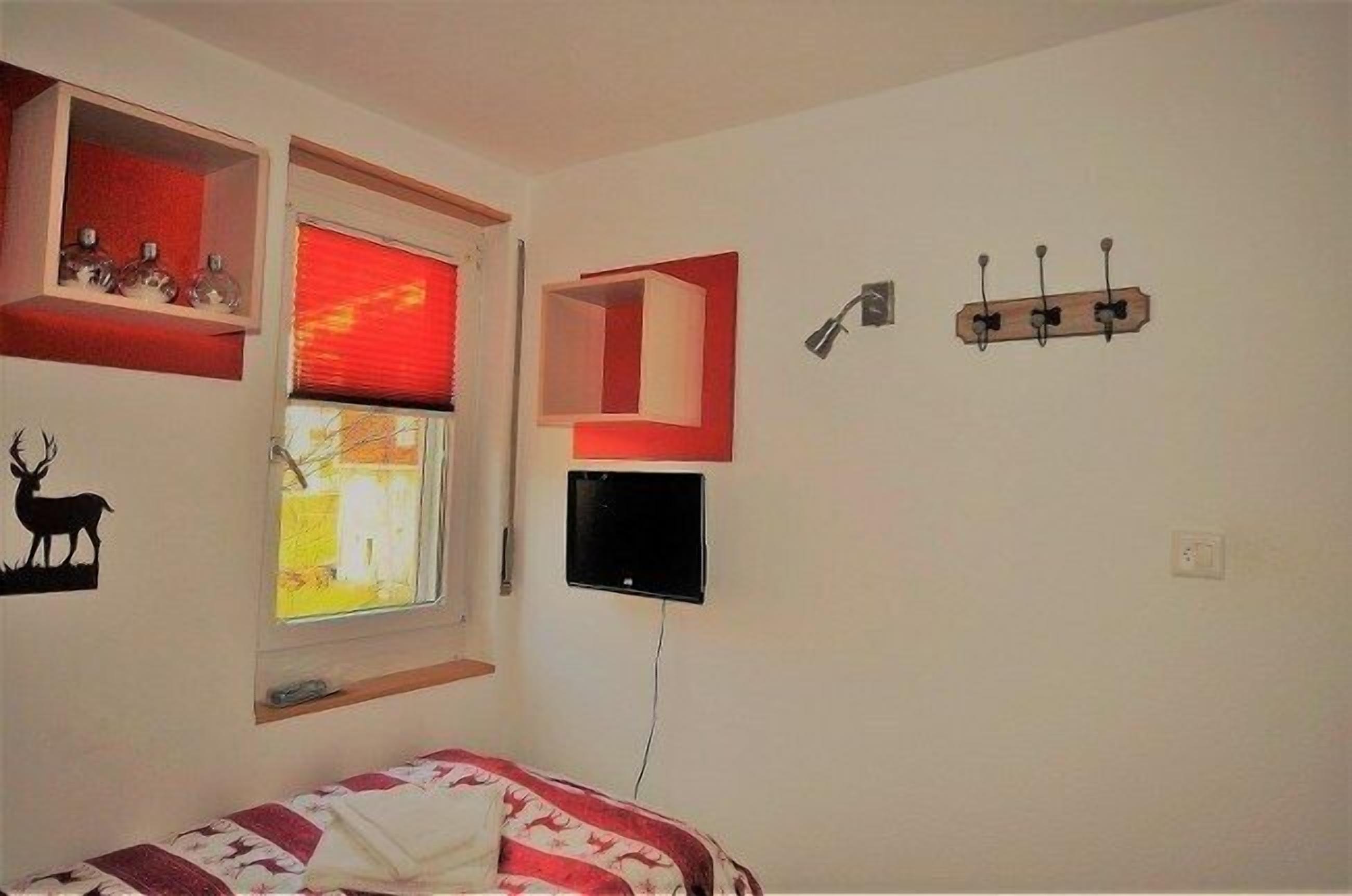 Un dormitorio con cama, ventana con persiana roja, televisor montado y ganchos en la pared.