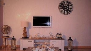 A living room with a white entertainment unit, a flat-screen TV, decorative items, and a clock on the wall. - Edelweiss Eden (Fiesch)