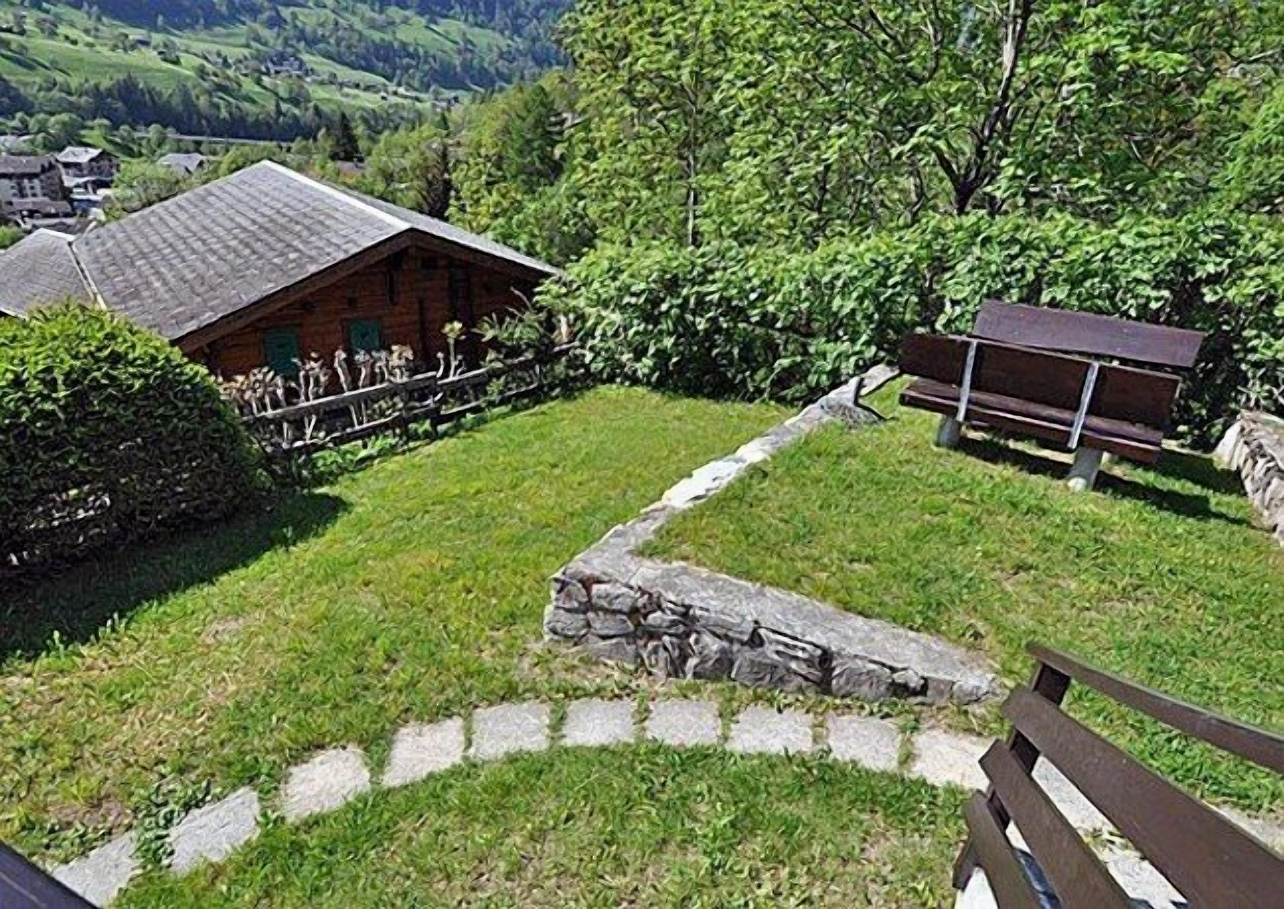 A rustic cabin with a stone foundation, surrounded by lush greenery and a stone wall.