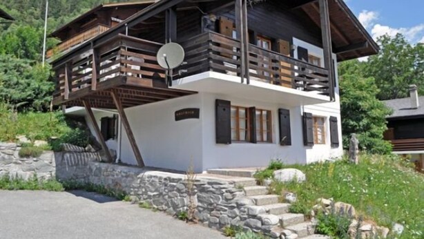 A two-story wooden chalet with a balcony, stone foundation, and a view of a mountainous landscape. - Melchior (Fiesch)