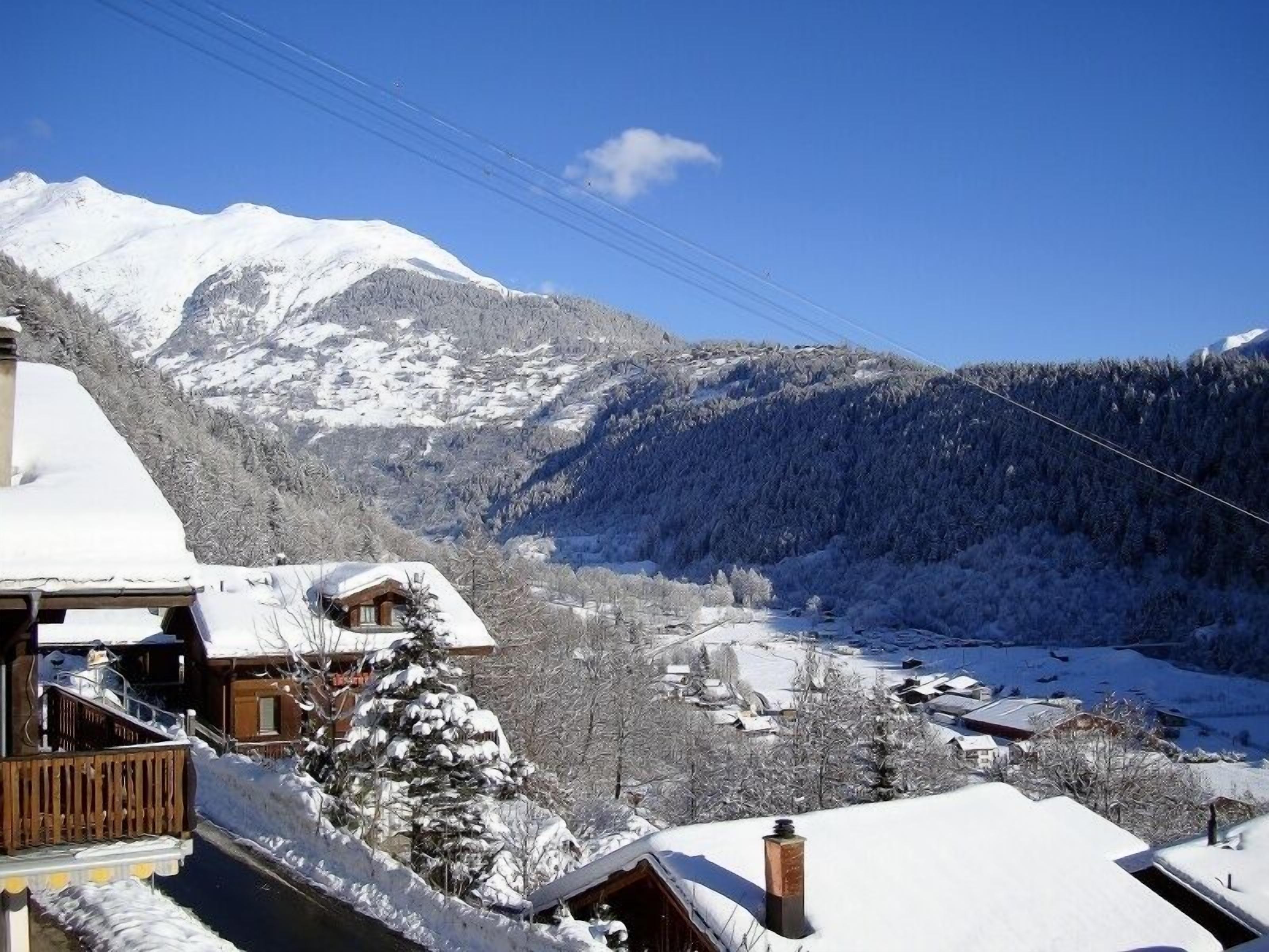 A mountain cabin with a wooden exterior and snow-covered roof, surrounded by a snowy landscape and a clear blue sky.