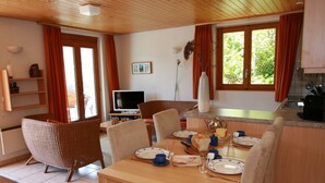 A dining area with a wooden table set for four, surrounded by wicker chairs. A television is mounted on the wall, and a window with curtains is visible. - Stachelbeere UG (Fiesch)