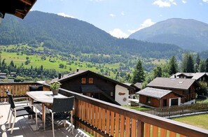 Una terraza de madera con una mesa y sillas, con vista a un paisaje montañoso con casas y una vía de tren.