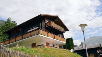 Un chalet de madera de dos pisos con balcones, fachada blanca y montañas al fondo.