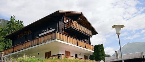 A two-story wooden chalet with balconies, a white facade, and a mountainous backdrop.