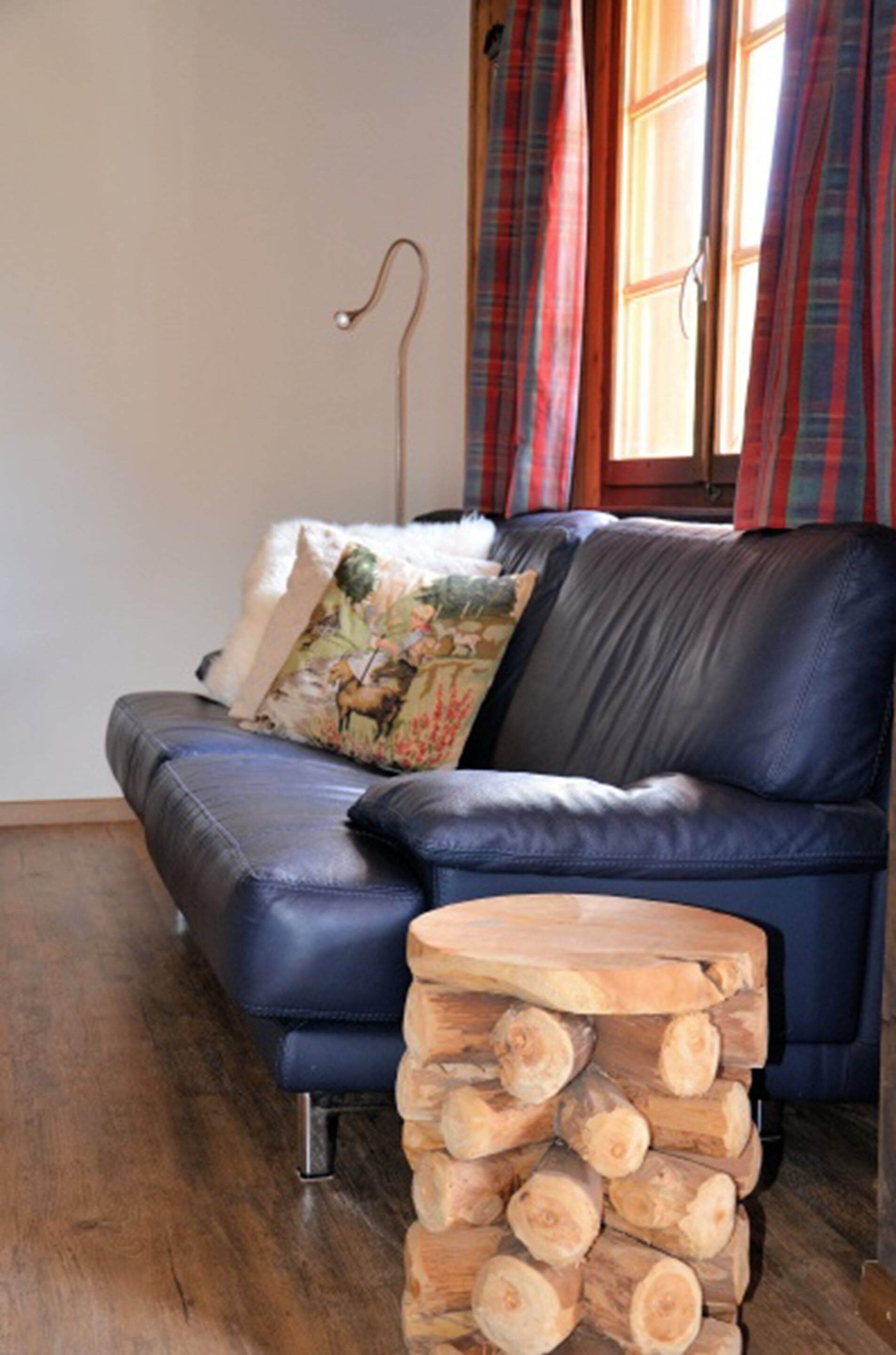 Un sofà blau de cuir amb coixins decoratius, una taula de fusta petita amb llenya i cortines de quadres a una finestra.