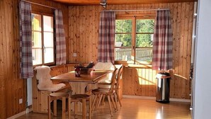 L’intérieur d’un chalet en bois comprend une table à manger, des chaises, un banc recouvert de fourrure, des rideaux à carreaux et une fenêtre donnant sur une terrasse.