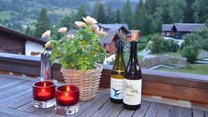 Une terrasse en bois avec un panier en osier contenant des fleurs blanches, deux bouteilles de vin et deux bougies allumées.