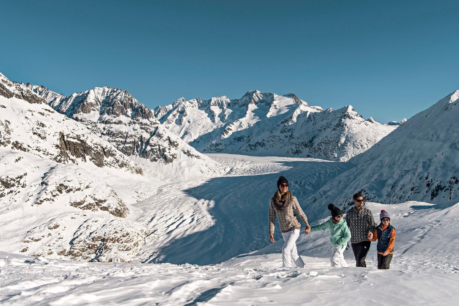 Eine Familie wandert auf einem verschneiten Bergpfad, im Hintergrund Gletscher und Berge.