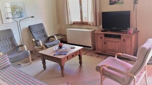 A living room with a sofa, two armchairs, a coffee table, a TV on a wooden cabinet, a radiator, and a window with curtains. - Wasserhügeli (Fiesch)
