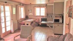 A cozy room with a wooden ceiling, a dining table with chairs, a sofa, a kitchen area with a microwave, and a window with curtains. - Wasserhügeli (Fiesch)