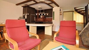 A modern living room with two red chairs, a wooden coffee table, and a kitchen area with a curved ceiling and pendant lights. - Wolf (Fieschertal)