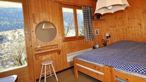 A wooden cabin bedroom with a bed, a round mirror, a stool, and a window overlooking a mountainous landscape. - Uf der Egga 419 (Fiesch)