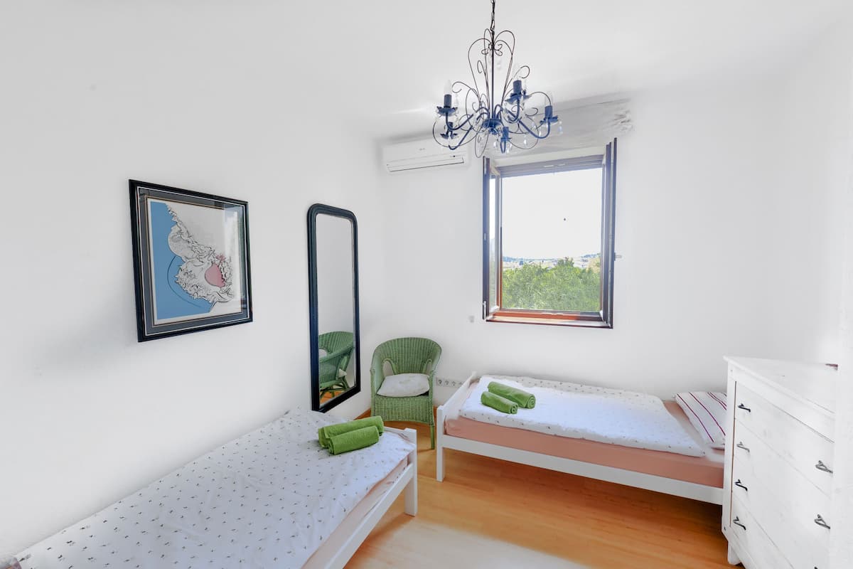 Un dormitorio con dos camas individuales, una silla verde, un aparador blanco, una araña, un mapa enmarcado y una ventana con vistas.
