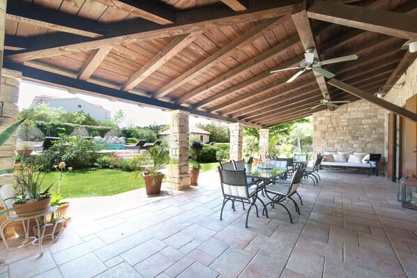 An outdoor patio with stone flooring, wooden beams, and a ceiling fan. There are potted plants and outdoor furniture. - Apartments Villa Renata (Murter)