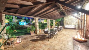 An outdoor patio area with stone flooring, wooden beams, a table with chairs, potted plants, and a stone wall in the background.