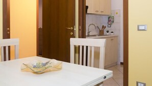 A room with a white table, a white chair, a wooden door, and a kitchen area with cabinets and a sink.