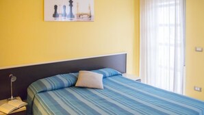 A bed with a blue striped bedspread and a pillow, a nightstand with a lamp and an open book, and a painting of chess pieces on the wall.