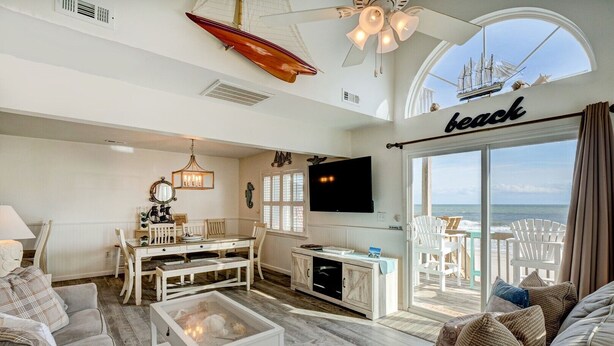 Un salón espacioso con un ventanal grande con vista a la playa, un comedor con mobiliario blanco y una zona de estar acogedora con chimenea.