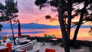 Een terras met tafels en stoelen, met uitzicht op een waterpartij en bergen tijdens zonsondergang.