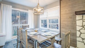 Un coin repas avec une table en bois dressée pour deux personnes, de grandes fenêtres avec des rideaux et un mur en pierre.