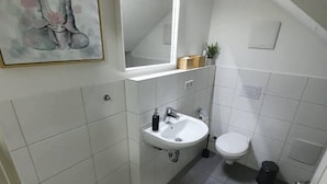 A small, clean bathroom with a white sink, toilet, and mirror. There is a small trash can and a potted plant on the counter. - White Pearl Hostel 2 (Nuremberg)