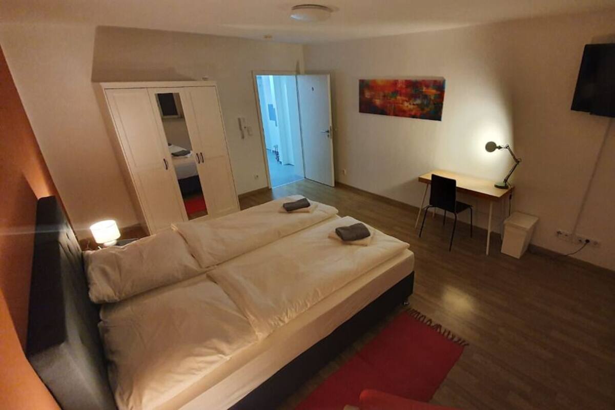 A bedroom with a bed, a desk, a TV, and a door leading to another room.