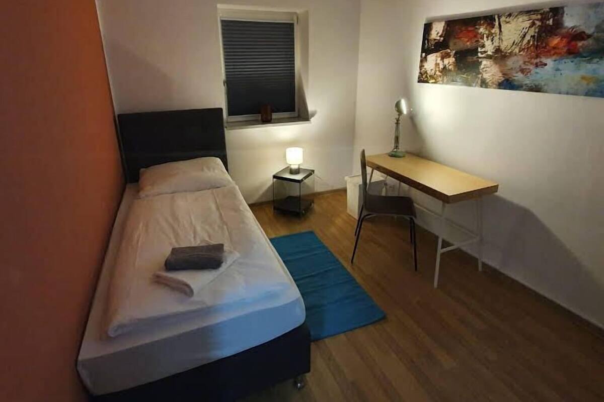 A small, tidy room with a bed, a desk, a chair, a window with blinds, and a painting on the wall.