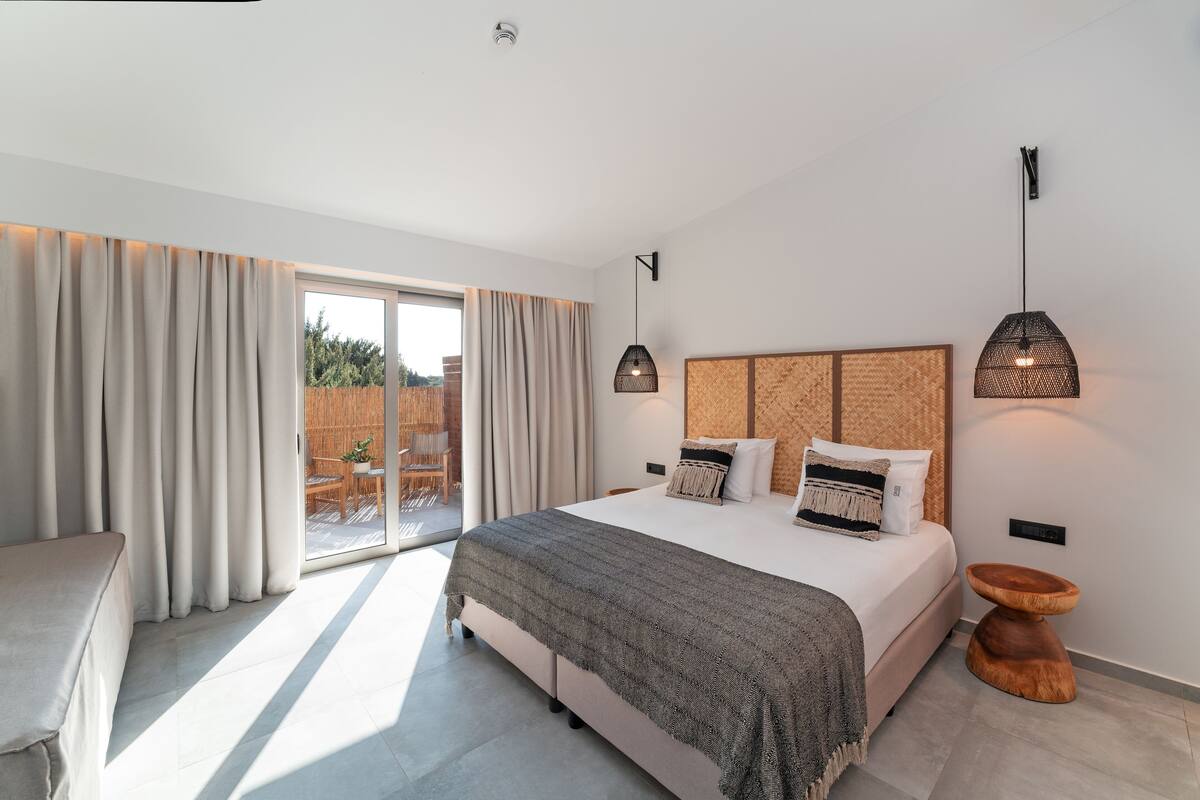 A modern bedroom with a large bed, a wooden side table, and a view of the outdoors through a sliding door.