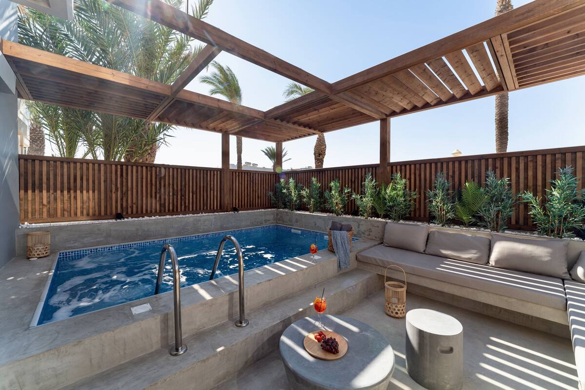 A rooftop pool area with a wooden pergola, a seating area with cushions, and a small table with a drink and grapes.