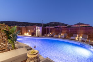 Piscina con sillas de descanso y sombrillas, rodeada de muros de madera y un paisaje montañoso.