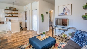 Una sala de estar moderna con sofá, reposapiés y una mesa de centro de vidrio. Hay una cocina con armarios blancos y encimera de madera. Un cuadro de un caballo está en la pared, y una planta en el suelo.