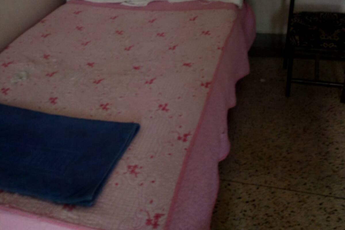 Un lit simple avec une couvre-lit rose et blanche, une couverture bleue et une chaise dotée d’une tête de lit sombre.