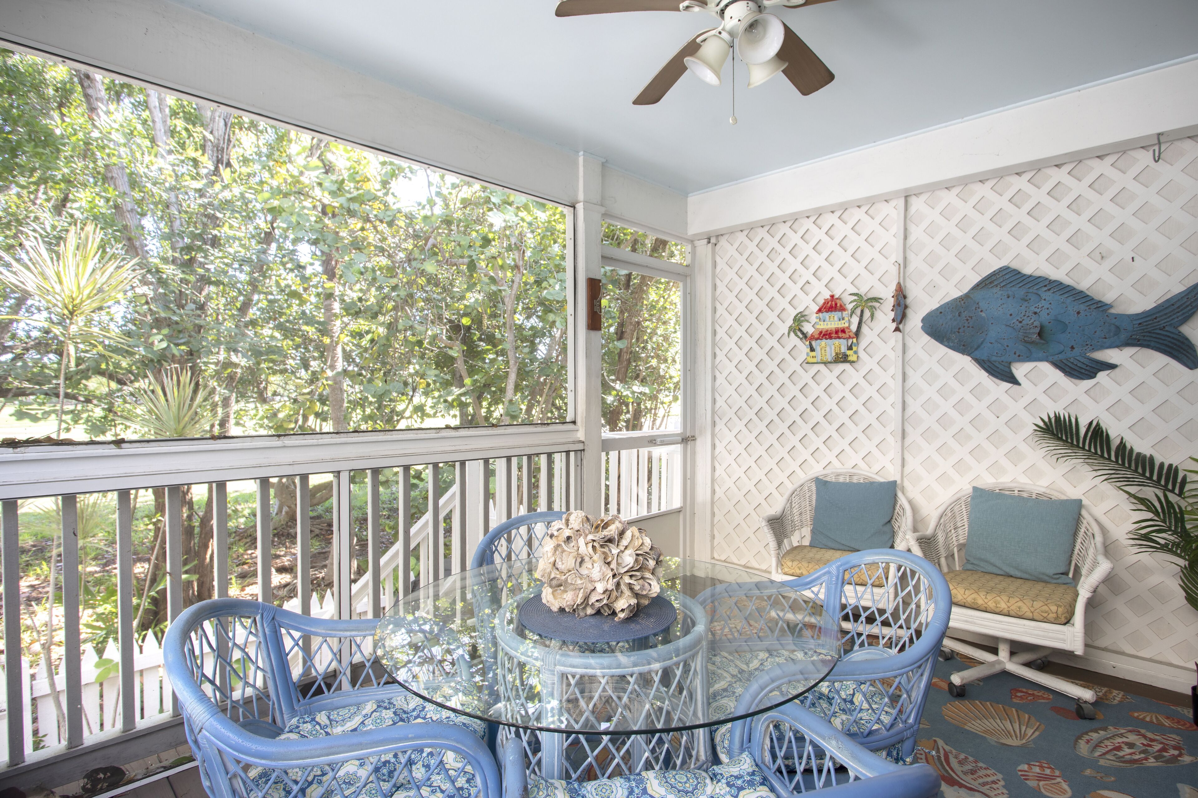 A glass-top table with blue chairs, a white lattice wall, and a ceiling fan.