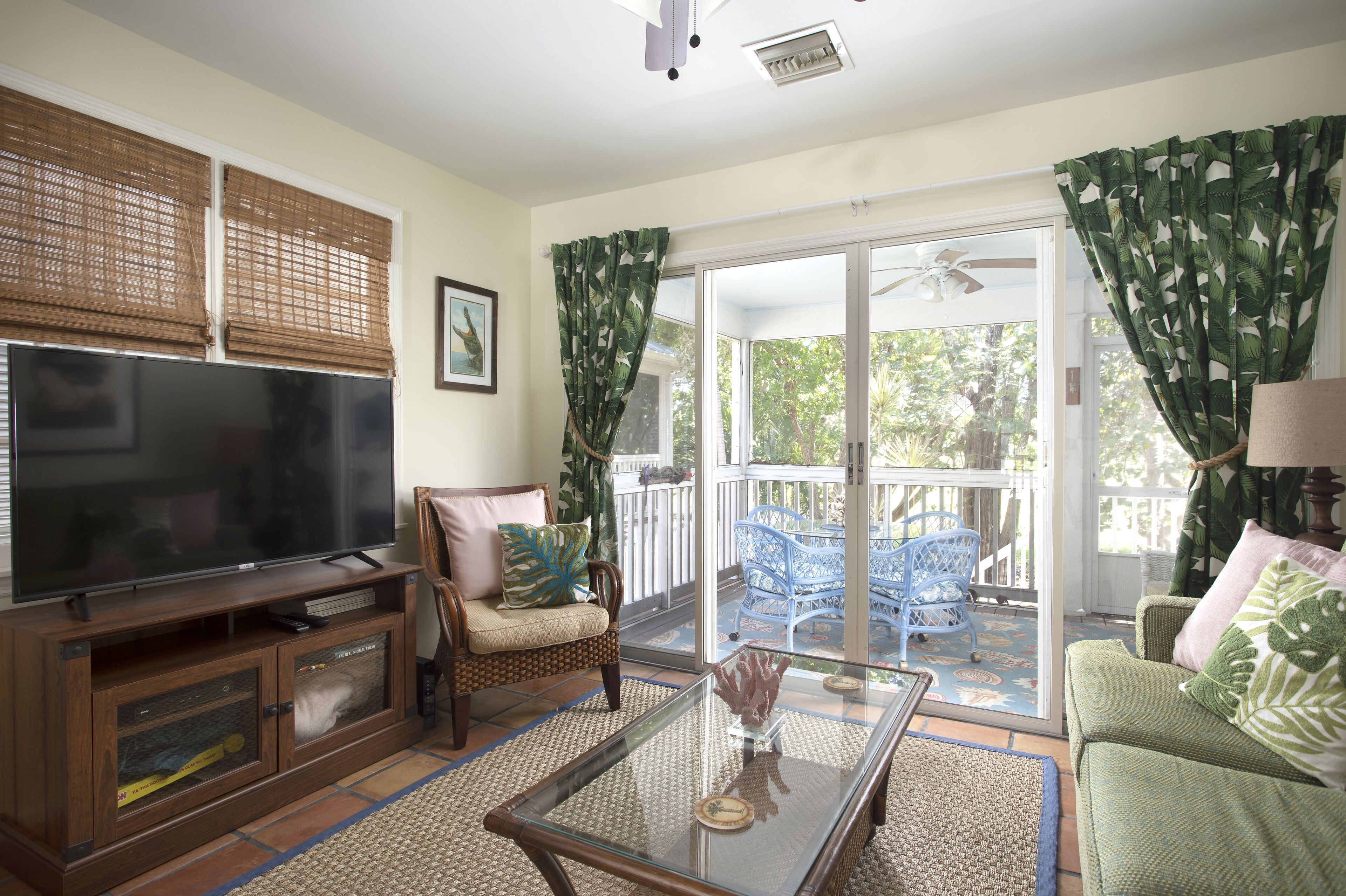 A living room with a TV, sofa, and chairs, a glass coffee table, and a sliding glass door to an outdoor patio.