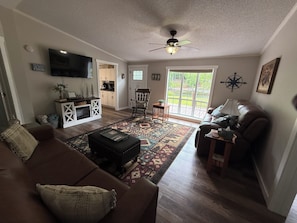 A living room with a brown sofa, a television, a ceiling fan, and a view of the outdoors through a sliding glass door.